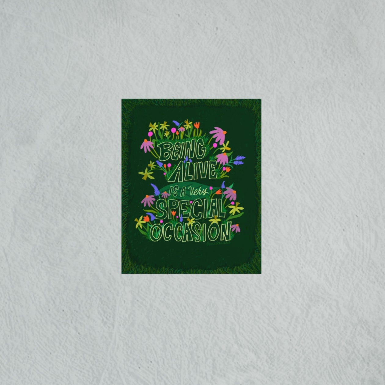 Decorative wall art with colorful text and flowers on a green background, placed on a textured white wall.